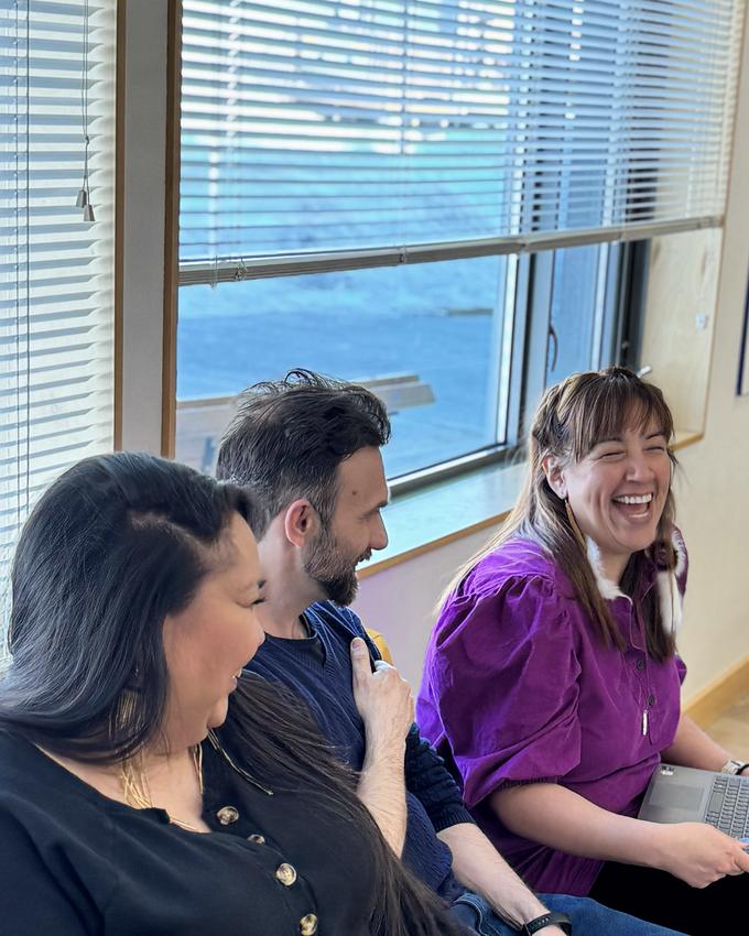 Three colleagues laughing together in a bright meeting room, one with a laptop open